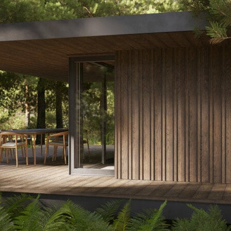 Brown Millboard Envello Siding on n a house tucked in the woods with a patio that has a table and chairs on it.