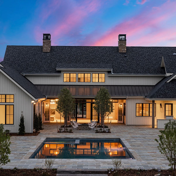 The back of this three story house has a elegant vibe with the the interior lights on and a pool that is all surrounded by outdoor patio space. 
