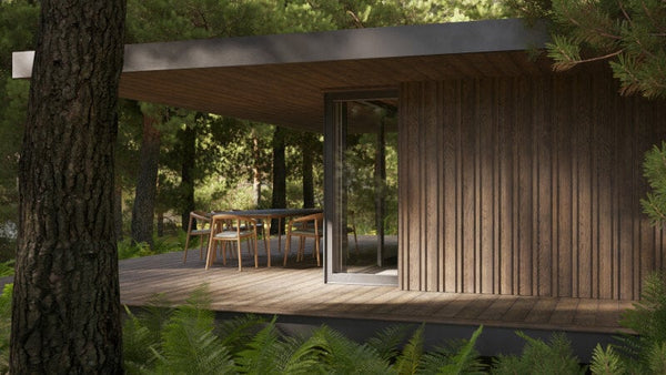Brown Millboard Envello Siding on n a house tucked in the woods with a patio that has a table and chairs on it.